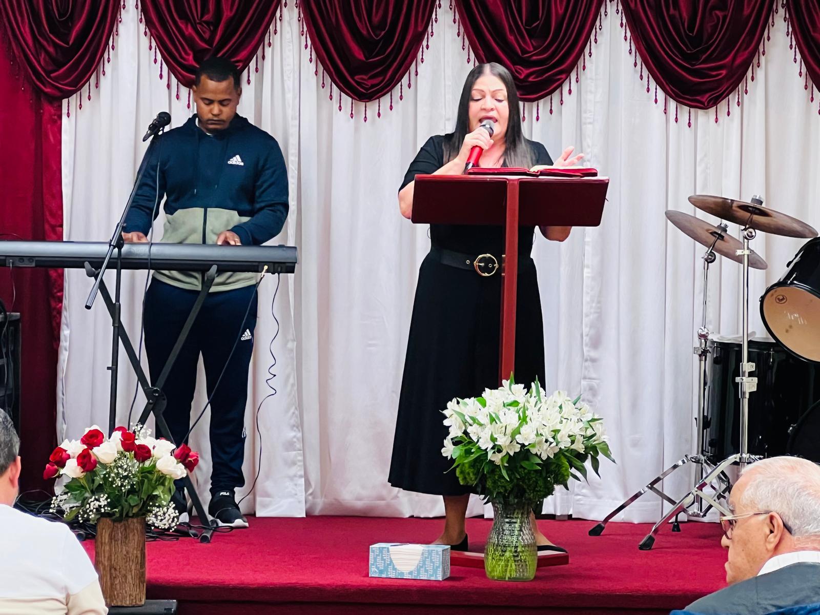 La pastora María Reyes predicando desde el púlpito, acompañada al teclado, durante un culto de la congregación